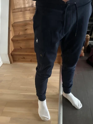 Mörkblå cargobyxor Polo Ralph Lauren - Mörkblå cargobyxor från Polo Ralph Lauren med snygga sidofickor med knapp, dragsko i midjan och broderad logga på benet. Tillverkade i mjuk bomullsmix för maximal komfort och avslappnad look. Perfekta för chill dagar eller streetwear. 