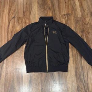 Svart vindjacka från EA7 Emporio Armani - Snygg svart vindjacka från EA7 Emporio Armani med guldfärgade detaljer och dragkedja. Jackan har ribbad krage och muddar, två fickor fram och meshfoder på insidan. Perfekt för dig som gillar stilrena och sportiga plagg med exklusiv känsla.