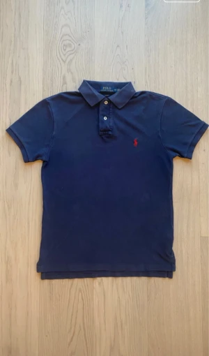 Mörkblå piké från Polo Ralph Lauren - Säljer en mörkblå pikétröja från Polo Ralph Lauren i slim fit. Klassisk design med krage, två knappar och röd broderad logga på bröstet. Tillverkad i 100% bomull för skön känsla. Perfekt för dig som gillar stilrena och tidlösa plagg.