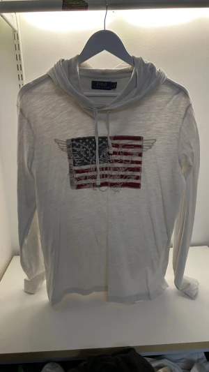 Vit hoodie Polo Ralph Lauren M - Vit långärmad hoodie från Polo Ralph Lauren i tunn bomull. Framsidan har ett tryck med amerikansk flagga och vingar, samt snörning i huvan. Perfekt för en chill och avslappnad stil.