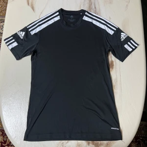 Svart Adidas t-shirt med vita ränder - Snygg svart t-shirt från Adidas med klassiska vita ränder på axlarna och logga på båda ärmarna. Modellen är kortärmad och tillverkad i mjukt, lätt polyester-material (Primegreen Aeroready) som andas. Perfekt för dig som gillar sportig stil.