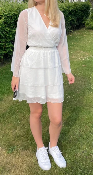 Vit omlottklänning med volang - Superfin vit kort omlottklänning med långa transparenta ärmar och volangdetaljer i kjolen. Klänningen har resår i midjan för en snygg passform och är gjord i ett lätt,  material som känns luftigt🤩 använd fåtal gånger🤍 säljer pga ingen användning🤍