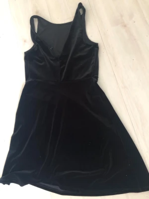 Svart sammet kortklänning - Supersnygg svart kortklänning i mjuk sammet med v-ringning och ärmlös design. Perfekt för dig som vill ha en enkel men elegant look. Klänningen har en lätt utställd kjol och smala axelband för en stilren vibe.