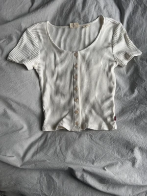 Vit ribbad topp med knappar från Levi's - Supersöt vit ribbad topp från Levi's med korta ärmar och snygg knappdetalj framtill. Modellen är croppad och har en rundad urringning. Perfekt att styla med jeans eller kjol för en chill look.