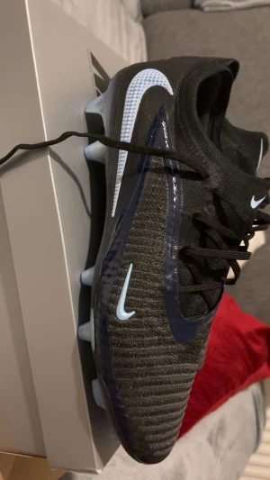 Nike Phantom GX 6 Elite Low FG fotbollsskor svart/blå - Nike Phantom GX 6 Elite Low FG fotbollsskor i svart och mörkblå med ljusblå detaljer och Swoosh-logga. Skorna har snörning, ribbad ovandel i syntetmaterial och platt sula med dobbar för gräsplan. Snygg och modern design som sticker ut på planen.
