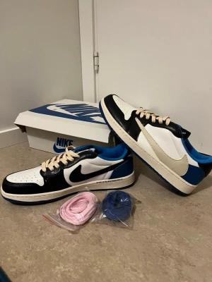 Nike Air Jordan 1 Low Travis Scott x Fragment - Nike Air Jordan 1 Low Travis Scott x Fragment med svart, vit och blå färgblockning. Swoosh-loggan är omvänd i beige, och skorna har krämfärgade snören samt extra snören i rosa och blått. Skinnmaterial och perforerad tå. Ikoniska detaljer som Cactus Jack och Air Jordan broderat på hälen.