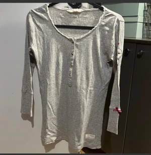 Grå ribbad långärmad tröja Odd Molly - Supersöt grå ribbad långärmad tröja från Odd Molly med knappar framtill, dekorativ vinröd rosett på ärmen och liten spetskant vid halsen. Skön och stretchig bomullskvalitet. Längd ca 60cm.