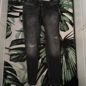 Svarta jeans med slitningar - Säljer ett par svarta jeans med coola slitningar och tvättad look. Modellen är skinny med smal passform och klassisk fyrficksdesign. Jeansen har låg midja och är gjorda i stretchigt denim för skön känsla. Perfekta för dig som gillar en edgy stil.