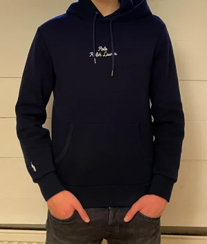 Marinblå hoodie från Polo Ralph Lauren - Snygg marinblå hoodie från Polo Ralph Lauren med broderad logga på bröstet och ärmen. Tröjan har huva och en stor magficka. Mjuk bomullskvalitet och långärmad passform, perfekt för chill dagar.