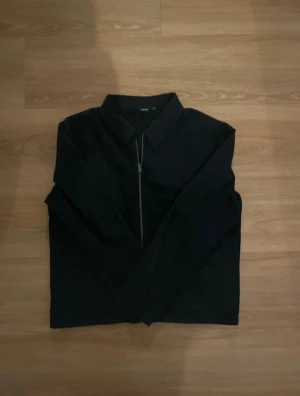 Svart overshirt från LMTD med dragkedja - Svart overshirt från LMTD i storlek M, med klassisk krage och silverfärgad dragkedja framtill. Jackan har raka långa ärmar och en enkel ficka på sidan. Materialet är bomull och ger en clean och stilren vibe.