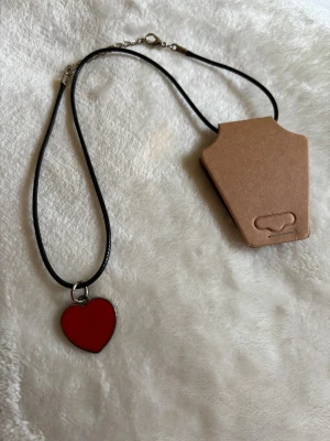 Halsband med rött hjärthänge - Svart halsband med ett rött hjärthänge i metall. Hängsmycket är blankt och har en enkel, stilren design. Kedjan är tunn och har ett klassiskt spänne. Perfekt accessoar för att addera lite färg och kärlek till din outfit.
