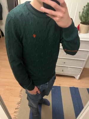 Mörkgrön stickad tröja med röd logga - Snygg mörkgrön stickad tröja från Morris med klassiskt kabelstickat mönster och rund hals. Tröjan har en röd broderad logga på bröstet och diskreta ribbade muddar vid ärmslut och nederkant. Perfekt för dig som gillar stilrena detaljer och skön passform. Storlek M däremot ingen tagg i nacken om storlek. Tröjan är i gott som nytt skick och passar mig som är 180 74kg
