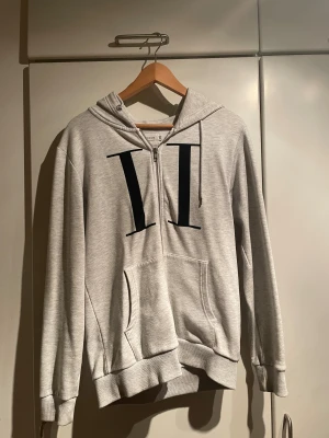 Grå zip-hoodie från Les Deux - Snygg ljusgrå hoodie från Les Deux med dragkedja, stor svart printad logga på bröstet och känguruficka. Mjuk insida och ribbade muddar. Klassisk stor huva. Perfekt för en chill och stilren look. Använd ett fåtal gånger.