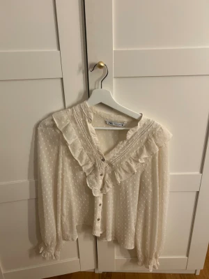 Vit blus med volanger från Zara XS - Supersöt vit blus från Zara i storlek XS med prickigt mönster, volangdetaljer längs halsringningen och knappar framtill. Blusen har transparent känsla, spetsinslag och långa ärmar med smock vid ärmslut. Perfekt för dig som gillar romantisk stil.