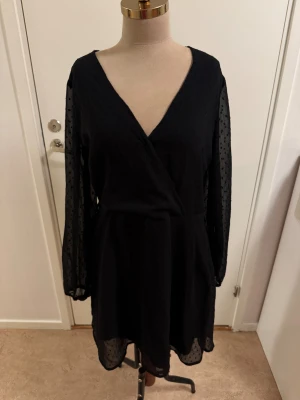 Svart omlottklänning Bubbleroom - Svart kort omlottklänning från Bubbleroom med transparenta långärmar i prickigt tyg. Klänningen har v-ringning och lätt utställd kjol. Materialet är mjukt och följsamt, perfekt för dig som gillar en stilren look med fina detaljer.