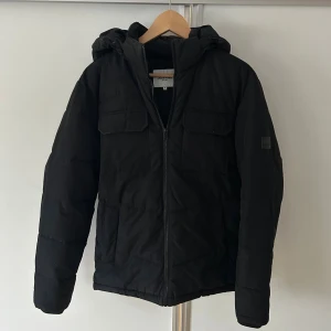 Svart pufferjacka med huva från Jack & Jones - Svart pufferjacka från Jack & Jones med huva, dragkedja framtill och två stora bröstfickor med lock. Jackan har quiltad design och är perfekt för kalla dagar. Snygg och enkel stil som funkar till det mesta. Jackan är i S men passar även mig som är medium nästan large 