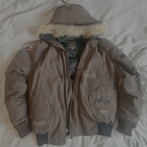 Brun Canada Goose dunjacka med päls - Säljer en brun dunjacka från Canada Goose med päls på luvan som ej går att ta bort. Jackan har broderade loggor av olika sponsorer (därav det lägre priset) , ribbade muddar och flera fickor med dragkedja. Insidan är svart och quiltad. (Säljer för att den är för stor för mig)