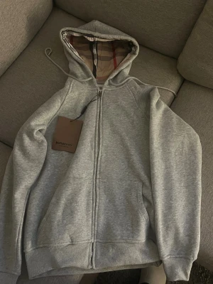 Grå hoodie från Burberry med dragkedja - Snygg grå hoodie från Burberry med klassiskt rutigt foder i huvan och broderad ryttare på bröstet. Tröjan har hel dragkedja, snörning i huvan och ribbade muddar. Perfekt för dig som vill ha en stilren och lyxig look med street-vibe. Den har inte varit använd alls
