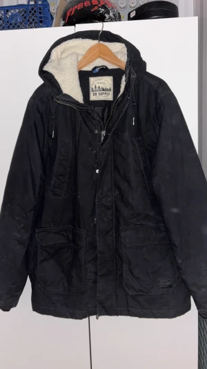 Svart parkajacka från DB Supply/H&M - Svart parkajacka från DB Supply med mysigt vitt teddyfoder i huvan. Jackan har dragkedja och knappar framtill, stora fickor och en klassisk parkamodell. Perfekt för kalla dagar när du vill hålla stilen och värmen.