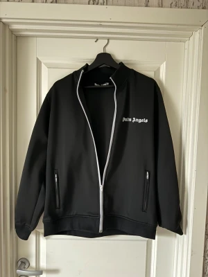 Palm Angels svart trackjacka - Palm Angels svart trackjacka med vita ränder längs ärmarna och vit logotyp på bröstet. Jackan har hel dragkedja framtill, två fickor med dragkedja och ribbade muddar. Materialet är mjuk polyester och passformen är loose.