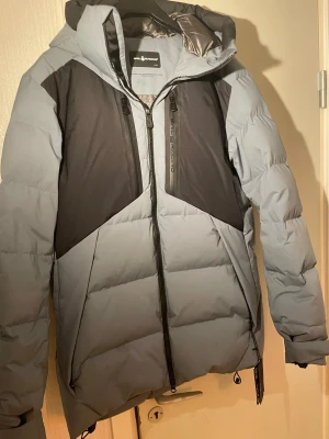 Grå dunjacka från Sail Racing - Snygg grå och svart dunjacka från Sail Racing med huva och flera dragkedjor framtill. Jackan har en quiltad design, vind- och vattenavvisande material samt certifierat dunfoder. Perfekt för kalla vinterdagar och har en modern, sportig look.