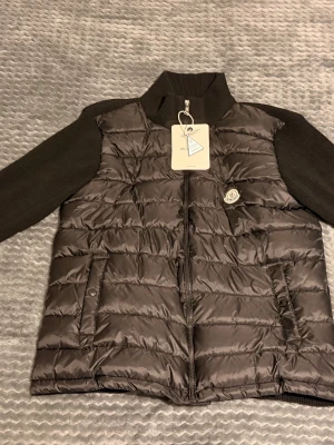 Moncler cardigan - Snygg brun dunväst från Moncler med quiltad framsida och ribbstickade ärmar och rygg. Västen har dragkedja framtill, två fickor med knapp och en hög krage. Klassisk Moncler-logga på bröstet och diskret broderad detalj på ärmen. Storlek S-M. Sitter enligt mig som en storlek M