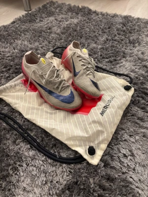 Nike Mercurial Air Zoom fotbollsskor - Säljer ett par Nike Mercurial Air Zoom fotbollsskor i vit med svart swoosh och röda detaljer. Skorna har snörning, spetsig tå och platt sula med dobbar. Materialet är syntet och mesh, med gula loggor på tungan och hälen. Perfekt för dig som vill ha grymma skor på planen. Använd i en säsong!