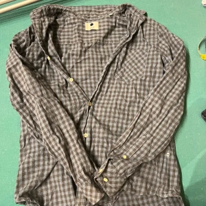 Rutig skjorta från NN07 - Säljer en slim fit skjorta från NN07 i storlek L. Skjortan har ett rutigt mönster i bruna och grå toner, klassisk krage, bröstficka och knappar framtill. Tillverkad i mjuk bomull som känns skön mot huden. Perfekt för dig som gillar stilren och avslappnad look.