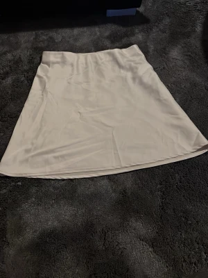 Ljusbeige satin-kjol från BikBok - Supersnygg ljusbeige kjol från BikBok i satinliknande material. Kjolen har en enkel och stilren design med lätt A-linjeformad siluett och resår i midjan för skön passform. Perfekt att styla med en crop top eller stickad tröja.