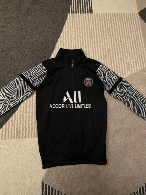 PSG träningströja med mönstrade ärmar - Säljer en svart PSG träningströja med coola svartvita mönstrade ärmar och halv dragkedja. Tröjan har PSG-logga på bröstet och Accor Live Limitless-tryck framtill. Perfekt för dig som gillar fotboll och vill ha en unik stil på träningen.