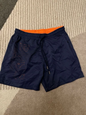 Mörkblå badshorts från BOSS - Säljer ett par mörkblå badshorts från BOSS med orange insida och snörning i midjan. Shortsen har ett stort BOSS-tryck på vänster ben och är tillverkade i syntetmaterial som torkar snabbt. Perfekta för strand eller poolhäng!