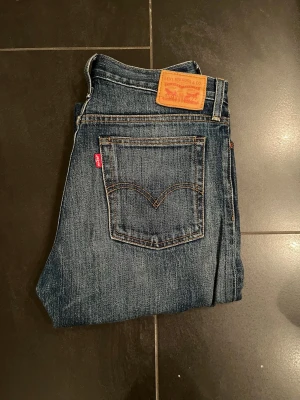 Levi's 501 blå jeans, storlek 28 - Klassiska Levi's 501 jeans i blå tvätt med raka ben och råa, fransiga benslut. Jeansen har knappgylf, fem fickor och den ikoniska läderlappen bak i midjan. Perfekt vintage vibe och snygg passform.