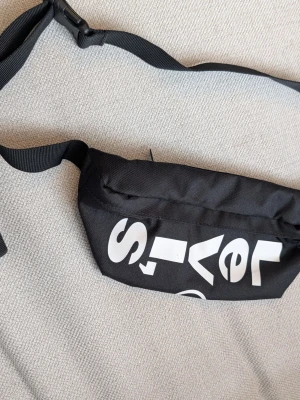 Svart magväska från Levi's - Säljer en svart magväska från Levi's med justerbart band och stor vit logga på framsidan. Väskan har dragkedja och är tillverkad i slitstark polyester. Perfekt för att bära det viktigaste när du är på språng.