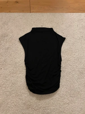Svart ärmlös topp från Gina Tricot - Svart ärmlös topp från Gina Tricot i mjukt material. Toppen har hög hals och snygga rynkade detaljer längs sidorna som ger en cool look. Perfekt att styla med jeans eller kjol för en trendig outfit.
