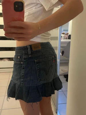 Levi's jeanskjol med volangkant - Supersnygg jeanskjol från gamla Levi's jeans med volangkant och råa kanter. Klassisk blå denim med femficksmodell och detaljerade sömmar. Sydd från gamla jeans men aldrig använd. Kjolen har normal passform och har midjemått 40 cm rakt över. 28 cm längd framtill i mitten och i mitten baktill 36cm.  Kjolen sitter lågmidjat på mig som brukar ha s/36.