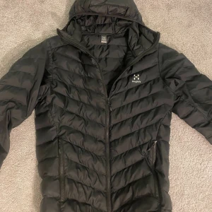 Svart pufferjacka från Haglöfs - Säljer en svart pufferjacka från Haglöfs med huva och dragkedja framtill. 