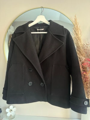 Svart skepparkavaj med stora slag - Säljer en svart skepparkavaj med dubbelknäppning och stora slag. Jackan har två stora fickor framtill och dekorativa knappar vid ärmsluten. Klassisk och tidlös modell i ullblandning, perfekt för kyligare dagar. Saknar en av knapparna och lite nopprig därav de billiga priset