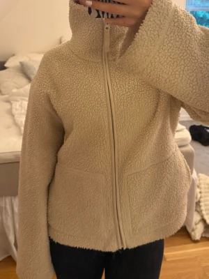 Beige teddyjacka med dragkedja - Mysig beige teddyjacka med hög krage och hel dragkedja framtill. Jackan har två stora fickor och är tillverkad i fluffigt teddy-material som håller dig varm. Perfekt för kyliga dagar och ger en avslappnad, trendig look.