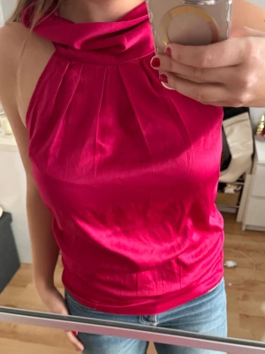 Hallonröd halterneck topp i satin - Säljer en hallonröd halterneck topp i glansigt satintyg. Toppen har hög krage med veckad detalj framtill och knäpps med två knappar i nacken. Öppen rygg med snygg cut-out och ärmlös design. Jätte snygg och skön!💕☺️