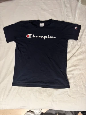Svart Champion t-shirt  - Säljer en svart t-shirt från Champion med stor vit logga och röd detalj på bröstet. Klassisk passform och korta ärmar. Ikonisk Champion-märke även på ärmen. Perfekt till streetwear-stilen.