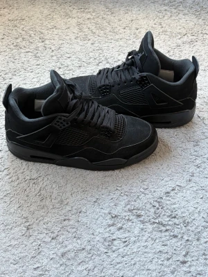 Jordan 4 black cat sneakers - Säljer ett par helsvarta Nike Air Jordan 4 sneakers med snygg mesh och mockadetaljer. Skorna har snörning, vadderad plös och ikoniska Jumpman-loggor på plös och häl. Tjock sula med synlig luftdämpning och coola plastdetaljer vid snörningen. Perfekta för dig som gillar streetwear.