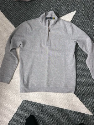Grå half zip tröja från Polo Ralph Lauren - Säljer en stilren grå långärmad tröja från Polo Ralph Lauren med half zip och liten broderad logga på bröstet. Tröjan är i mjuk bomull och har en hög krage som ger en clean look. Perfekt till lager på lager under höst och vinter.