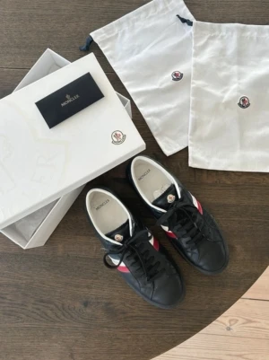 Moncler svarta sneakers med röd/vit detalj - Snygga svarta sneakers från Moncler med klassiska rödvita ränder på sidan och logga på plösen. Skorna har svarta snören, vit insida och är tillverkade i skinn. Perfekta för dig som vill ha en clean och sportig look med exklusiv känsla.