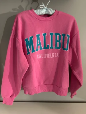 Rosa sweatshirt med Malibu-tryck - Säljer en rosa sweatshirt med stort turkost och vitt 'MALIBU CALIFORNIA'-tryck på bröstet. Tröjan har rund hals, ribbade muddar och en relaxed passform. Perfekt för dig som gillar färg och vill ha en chill college-vibe.