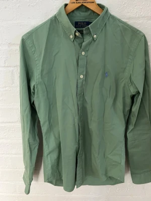 Grön skjorta från Polo Ralph Lauren - Snygg grön skjorta från Polo Ralph Lauren i slim fit med klassisk button-down krage och broderad logga på bröstet. Tillverkad i lätt bomullstyg som känns skönt mot huden. Perfekt till jeans eller chinos för en clean look.
