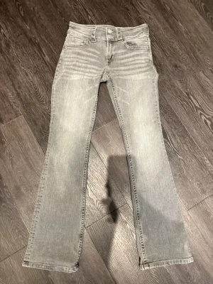 Grå bootcut jeans med detaljer - Säljer ett par grå jeans med bootcut passform och snygga detaljer som knappar på bakfickorna och extra bälteshällor. Jeansen har en ljus tvätt och raka ben med lätt utsvängning. Materialet är klassisk jeans och de har en modern look.
