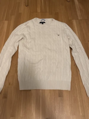 Vit kabelstickad tröja från Gant - Kan gå ner i pris ordentligt! Kabelstickad vit tröja från Gant med rund halsringning och diskret logga broderad på bröstet. Tröjan har långa ärmar och ribbade muddar vid ärmslut och nederkant. Perfekt för dig som gillar klassisk och clean stil.