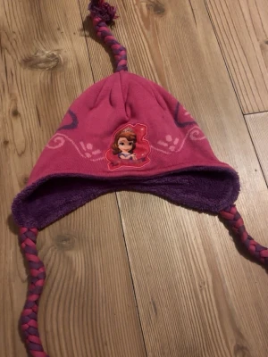 Rosa och lila mössa med flätor - Söt mössa i rosa och lila med flätade snören och fleecefoder. Mössan har ett tryck framtill med prinsessmotiv och dekorativa mönster i rosa och lila. Perfekt för kalla dagar och håller öronen varma.