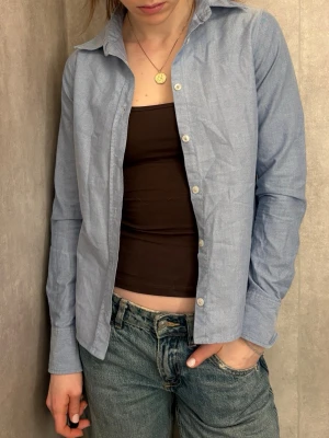 Ljusblå klassisk skjorta - Ljusblå skjorta i bomull med klassisk krage och långa ärmar. Skjortan har normal passform och stängs med vita knappar framtill. Perfekt att styla öppen över ett linne eller knäppt för en clean look.