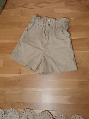 Beiga högmidjade shorts - Säljer ett par beiga shorts med hög midja och resårkant. De har två fickor fram, dubbla silverknappar och är gjorda i 100% bomull. 